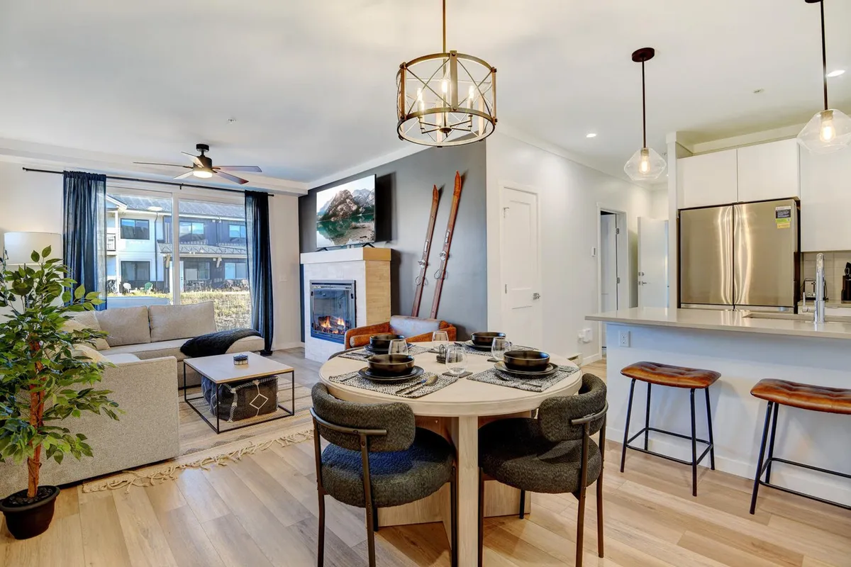 The image showcases a cozy and stylish open-concept living area featuring a comfortable sofa, an inviting fireplace, a chic dining table set for guests, and large windows that frame breathtaking views of the Colorado landscape. Decorative elements and soft lighting create a welcoming atmosphere, perfect for relaxing or gathering with loved ones after a day of outdoor adventures.