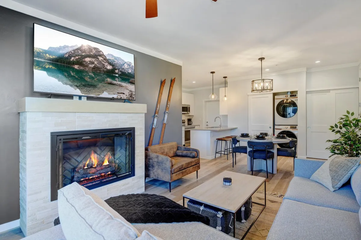 An open-concept space combining a cozy living area with a plush sofa, a wall-mounted TV above a color-changing electric fireplace, and an adjacent dining area with a wooden table and chairs. Large windows offer mountain views, creating a bright and inviting atmosphere.