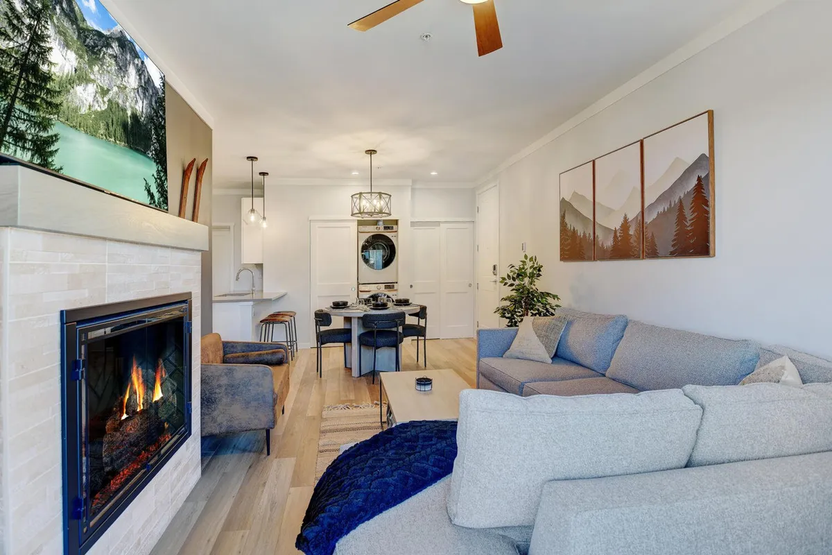 A warm and comforting living and dining space featuring a stylish fireplace, comfortable sofa, dining table with seating for four, mountain-themed artwork, and a pendant light. The room is ideal for both relaxing and hosting intimate dinners.