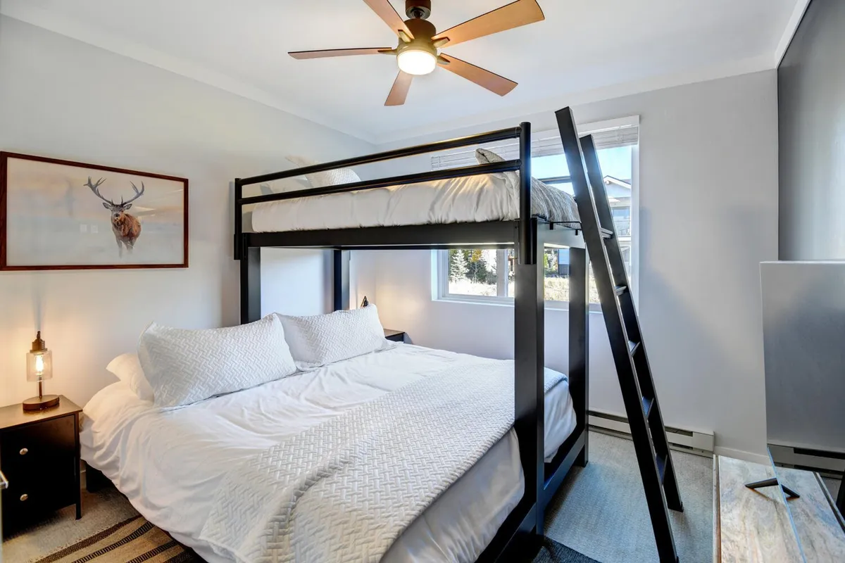 This image showcases a cozy bedroom featuring a twin-over-king bunk bed, a bedside lamp that offers warm lighting, framed artwork on the wall, and a window letting in gentle natural light. The playful yet restful atmosphere is perfect for families or groups with kids, inviting bedtime stories and memorable mornings.