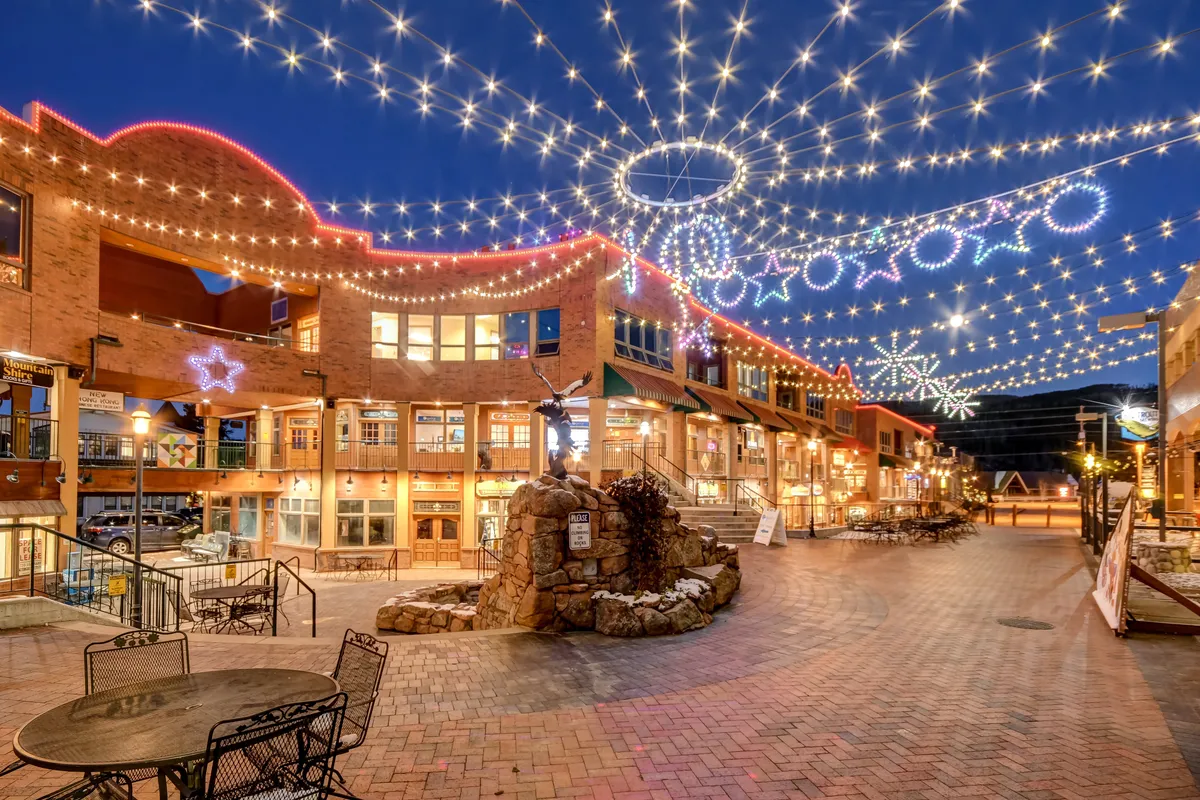 A lively exterior shot of a shopping and dining area illuminated by decorative string lights, with a stone fountain and inviting seating arrangements. The setting exudes a festive, social atmosphere, hinting at the vibrant downtown dining and entertainment scene available to guests.