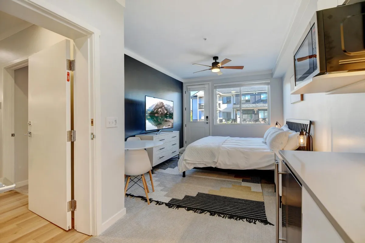 A spacious and modern master bedroom featuring a plush bed, large windows that flood the room with morning light, a dresser, a comfortable chair, and a television. The room exudes comfort and privacy, ideal for restful nights and leisurely mornings.
