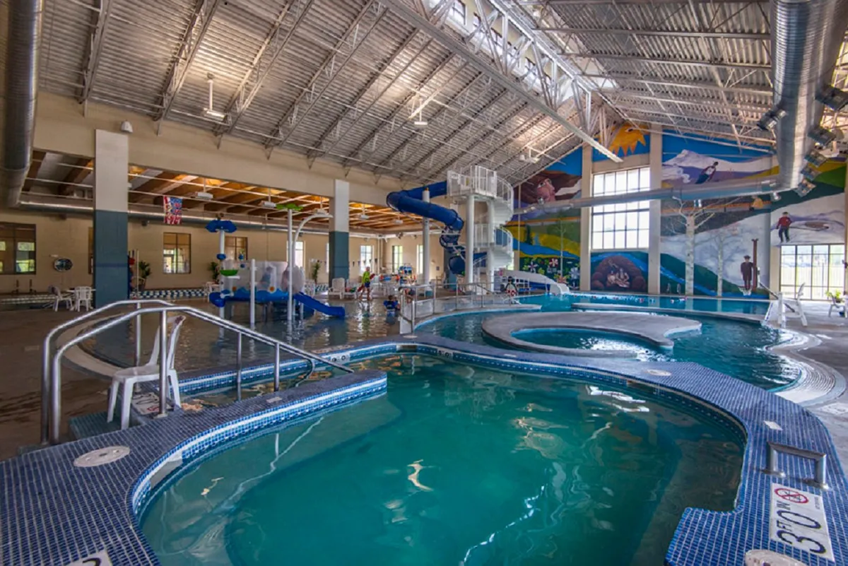 An energetic indoor waterpark scene with children playing in the pool and on colorful water slides. The space is filled with natural light and vibrant murals, creating a joyful and lively atmosphere.
