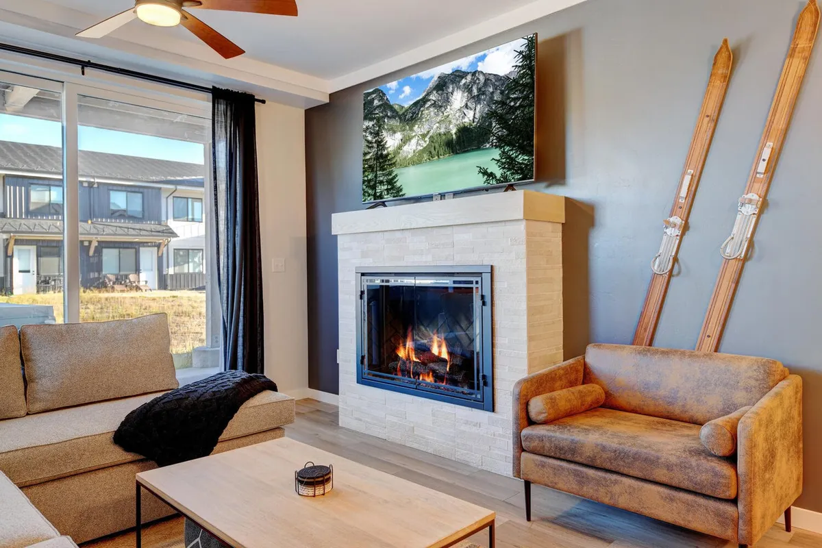 A cozy living area featuring a fireplace, TV, sofa, armchair, and skis leaned in the corner. The ambiance is warm and inviting, ideal for relaxing after a day of outdoor adventures. The visible elements hint at the condo's readiness for both winter and pet-friendly living.